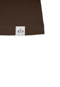 Ruskea puuvillakangas, jossa on taitettu reuna; vasemmassa alakulmassa on valkoinen etiketti, jossa lukee "BR" ristikkäin olevien välineiden kanssa.