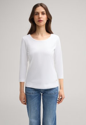 Weißes, langärmeliges Hemd mit rundem Ausschnitt und glatter Textur. Mit subtiler Logoschrift am Saum, kombiniert mit hellblauen Denim-Jeans.