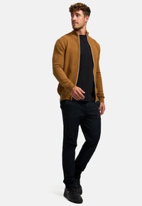 Brun texturerad zip-up cardigan med ribbat mönster, matchad med en svart t-shirt och mörka byxor. Modellen står framför en ljus bakgrund.