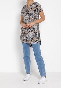 Kvinde iført en kortærmet paisley-tunika, lyseblå jeans og hvide sneakers, der står mod en ensfarvet hvid baggrund.