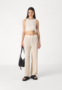 Beež cropped tank top, pikad kudumisega püksid vertikaalse õhulise mustriga ja must lahtine käekott. Modell kannab musti liugjalatseid.