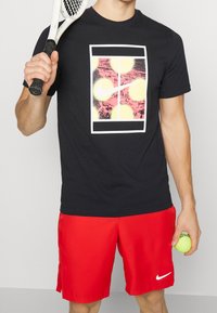 Camiseta negra con un estampado gráfico vibrante que presenta círculos amarillos y rosas. Combinada con pantalones cortos rojos, sosteniendo una raqueta de tenis y una pelota.