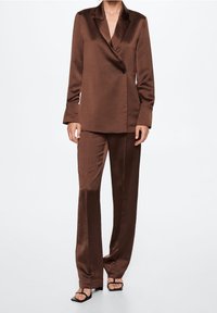 Hnědý saténový oblek skládající se z přizpůsobeného saka s jedním knoflíkem a kalhot rovného střihu, doplněný černými sandály na pásku.