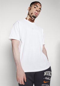 Biela bavlnená tričko s krátkymi rukávmi, s malým vyšitým logom Nike v rovnakých farbách na hrudi.