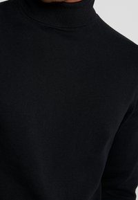 Suéter de cuello alto negro fabricado con tejido de punto acanalado, que presenta un diseño ajustado y una textura suave. Realza la zona del cuello.