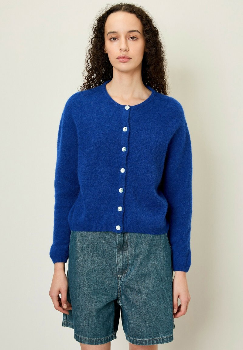 Blauwe, fuzzy gebreide cardigan met een ronde halslijn, lange mouwen en witte knopen aan de voorkant, gecombineerd met denim shorts.