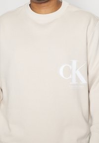 Beige Sweatshirt aus weichem Stoff, mit einem weißen Calvin Klein Logo und Text auf der Vorderseite. Rundhals und gerippte Bündchen.