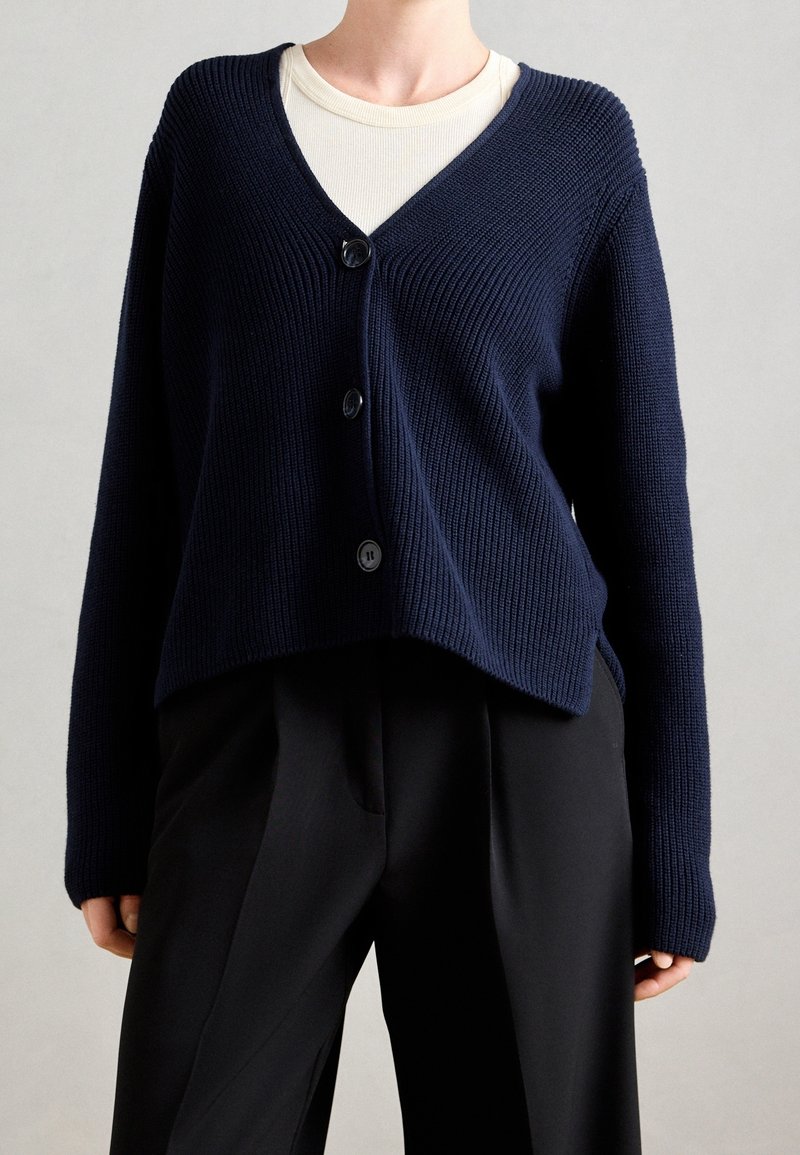 Cardigan en maille marine avec un col en V, des manches longues et trois boutons noirs. Porté sur un haut beige clair et assorti à un pantalon noir.