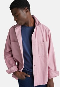 Růžová bavlněná košile s dlouhými rukávy, zapínáním na knoflíky, se dvěma náprsnými kapsami a stojáčkem, nošená přes tmavě modré tričko.