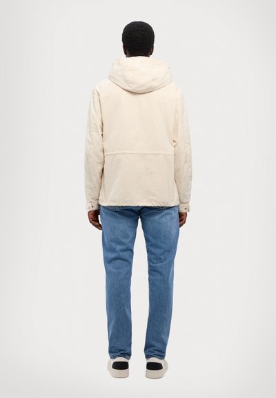 Bežna jakna s kapuco, ravnega kroja, z detajlom šiva na hrbtni strani, v kombinaciji z svetlo modrimi kavbojkami in čevlji z črnimi detajli.