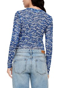 Langsleeve, aangesloten top van transparant blauw materiaal met een golvende camouflagepatroon; gecombineerd met middelmatige wassing, high-waisted jeans met achterzakken.
