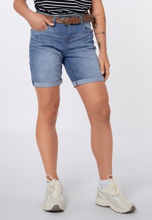 Persoon draagt opgerolde spijkershorts, gevlochten bruine riem, gestreept zwart-wit shirt, witte sneakers en witte enkelsokken, staat met één hand in de zak.