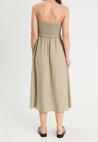 Vestido midi color caqui con cuerpo fruncido, tirantes delgados ajustables y tejido fluido. Combinado con sandalias planas, mostrando un corte holgado y caída.