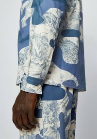 Miehen käsi lepää vartalon vieressä, päällä sinivalkoinen abstrakti kuviopaita ja housut, joissa on yksityiskohtaisia maisemaprinttejä.