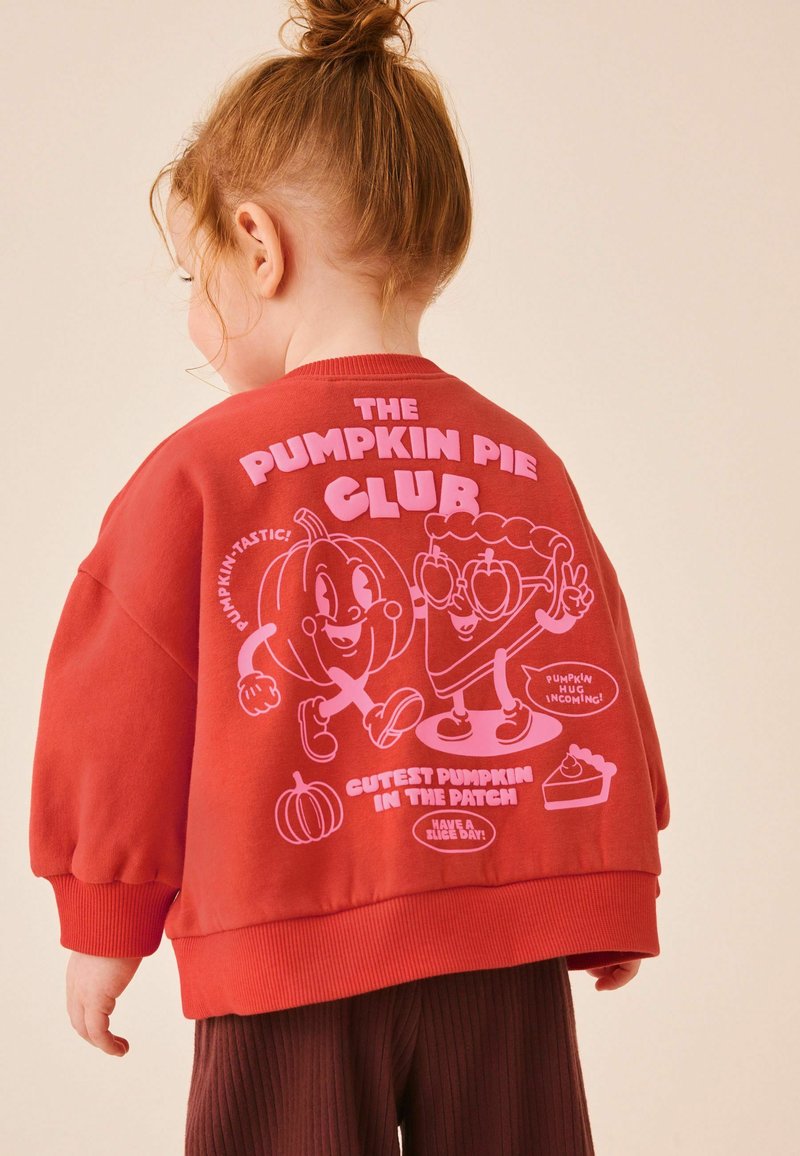 Niño pequeño con cabello rojo recogido en un moño que lleva una sudadera roja con una calabaza de dibujos animados y pastel con texto "The Pumpkin Pie Club" en la espalda.