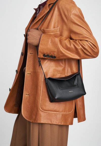 Bolso cruzado de cuero negro con un acabado texturizado, que presenta un diseño de solapa simple y una correa delgada ajustable, llevado sobre un blazer de cuero marrón.