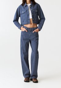 Femme portant une veste courte à rayures verticales bleu et blanc et un pantalon assorti, avec des baskets marron et noires, les mains dans les poches.