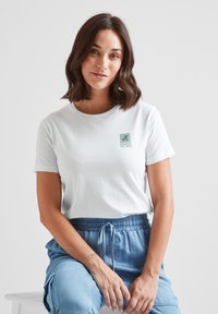 White cotton t-shirt with short sleeves, featuring a green "Respect the Wild" logo. Model wears blue drawstring pants, seated on a white bench.