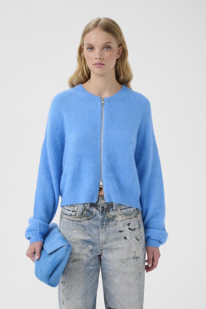 Heleroheline kampsun ümmarguse kaelusega, esil tõmblukuga ja mansettidega varrukatega. Kandmisega on seotud kulunud helesinised teksad ja sobiv sinine käekott.