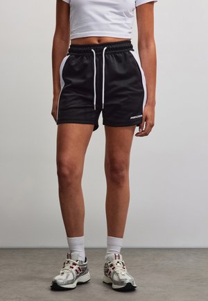 Person iført sorte sportsshorts med hvide sidelister, hvide snore, hvide ankelsokker og hvide sportssko, der står på gråt gulv.