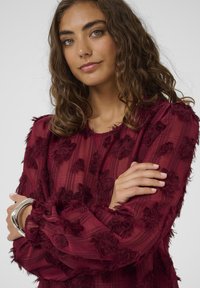 Blusa bordeaux realizzata in tessuto trasparente e strutturato con decorazioni floreali e scollatura rotonda. Maniche lunghe con dettagli sfrangiati.