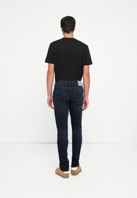 Sort kortærmet t-shirt parret med tætsiddende mørkeblå jeans. Jeansene har baglommer og en grå taljebåndlabel. Brune sko fuldender looket.