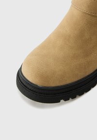 Bota de ante marrón con punta redondeada y suela de goma negra. La suela presenta una superficie rugosa para mayor tracción. Detalles de costura a lo largo de la parte superior.