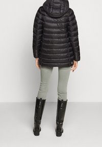 Černá péřová bunda s kapucí a prošívaným vzorem, doplněná světle zelenými úzkými kalhotami a černými gumovými botami nad kolena.