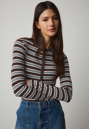 Mladá žena s dlhými hnedými vlasmi, ktorá nosí hnedo-bielo-modré pruhované tričko s dlhým rukávom a vysokými džínsami, s prekríženými pažami.