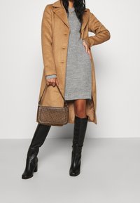 Cappotto lungo in lana color cammello con colletto a punta, abito a maglia grigio aderente, stivali neri fino al ginocchio e una borsa a tracolla con motivo e manico corto.