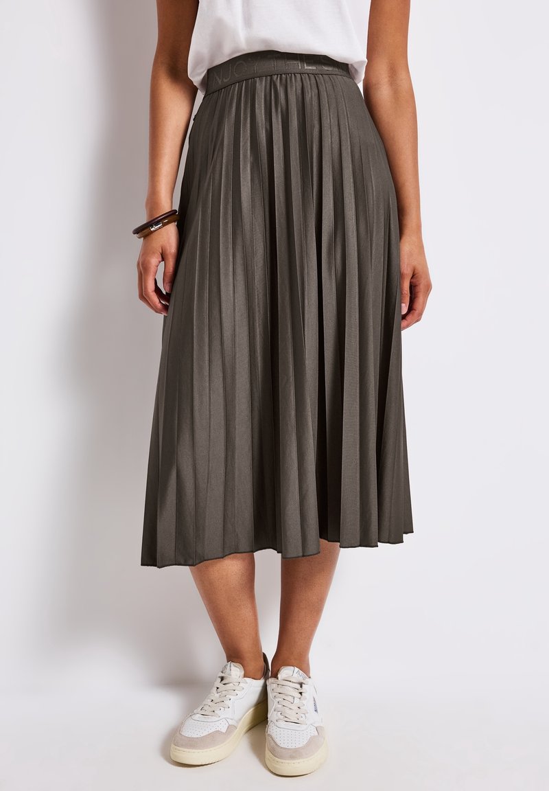 Femme portant une jupe midi plissée gris foncé, un haut blanc, des baskets blanches et des bracelets foncés au poignet gauche, debout devant un fonds uni.
