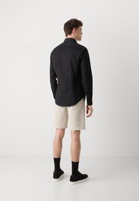 Black long-sleeve shirt with a collared neck, worn with beige shorts and black shoes. Minimalist design, smooth texture.