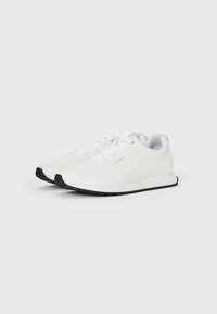Une paire de baskets blanches basse à semelles noires, avec un laçage classique et un logo discret sur le côté, présentée sur un fond clair uni.