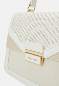 Vitt vävt handtag med en strukturerad design, texturerad topp, beige detaljer, guldfärgad hårdvara och en graverad varumärkesplakett. Ficka med lock.