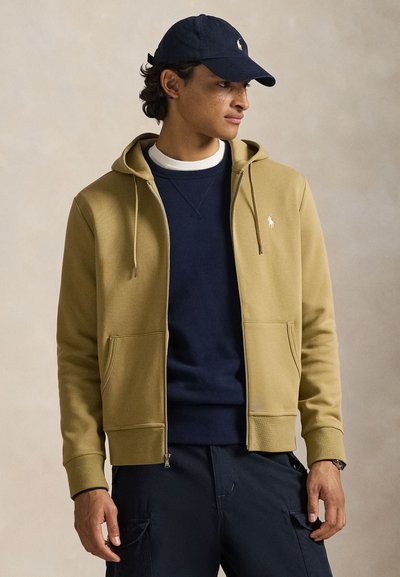 Φούτερ με κουκούλα και φερμουάρ σε μπεζ μείγμα βαμβακιού· διαθέτει μπροστινές τσέπες και ένα μικρό λογότυπο. Φοριέται πάνω από μπλε σκουρόχρωμο πουλόβερ.