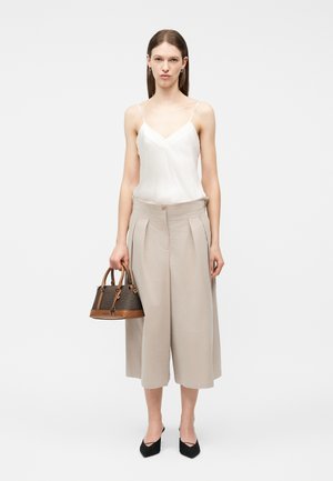 Γυναίκα που φοράει λευκό αμάνικο τοπ και μπεζ παντελόνες culottes, κρατώντας καφέ τσάντα, μαύρα μυτερά παπούτσια, όρθια μπροστά σε απλό φόντο.