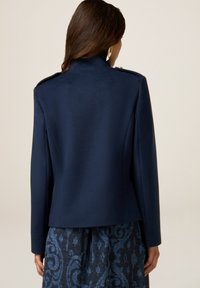 Marineblaue, taillierte Jacke aus strukturiertem Stoff mit hohem Kragen und Schulterklappen, kombiniert mit einem gemusterten blauen Rock. Rückansicht.