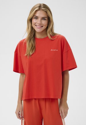 Young woman with long blonde hair smiles, wearing a loose red t-shirt and matching red pants with beige side stripes.