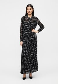 Μαύρο φόρεμα με λευκά πουά και συνοδευτικό μακρυμάνικο μπλουζάκι. Διαφανές ύφασμα, πλισέ φούστα, λεπτομέρεια με κορδόνι στη λαιμόκοψη, συνδυασμένο με μυτερές μαύρες γόβες.