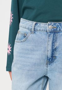 Jean en denim bleu clair avec un bouton à l'avant et des poches, porté avec une chemise verte à manches longues ornée de motifs de soleil roses sur les manches.