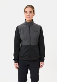 Vrouw in een zwart en grijs zip-up jack met logo op de borst en zwarte broek, staand tegen een effen witte achtergrond.