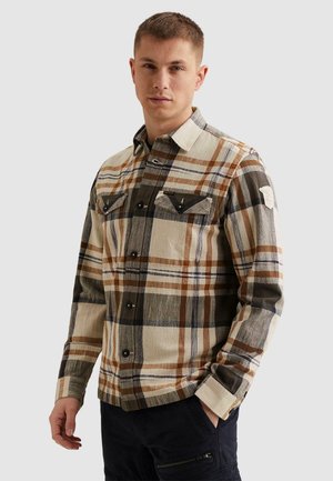 Mladi moški s kratkimi lasmi, oblečen v bež, rjavo in črno karo srajco s sponkami ter temne hlače, stoji z eno roko v žepu.