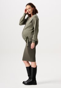 Olivgrüne Strickjacke mit einem Bindegürtel in der Taille, kombiniert mit einem gerippten olivgrünen Rock und schwarzen Stiefeln bis zum Knie. Glatte Textur.