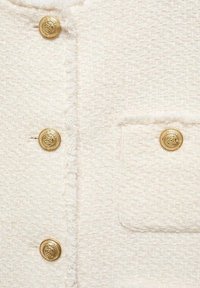 Chaqueta de tejido blanco texturizado con botones dorados y un bolsillo, con un borde deshilachado a lo largo de las costuras para un detalle adicional.
