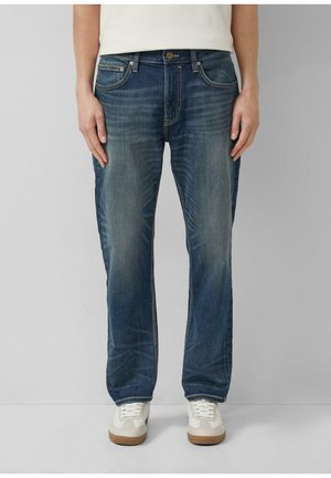 Mann trägt ausgewaschene blaue Jeans und weiße Turnschuhe, steht vor einem schlichten hellgrauen Hintergrund.