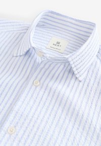 Chemise à manches courtes rayée bleu clair et blanc avec col boutonné, portant une étiquette "NEXT" et trois boutons à l'avant.