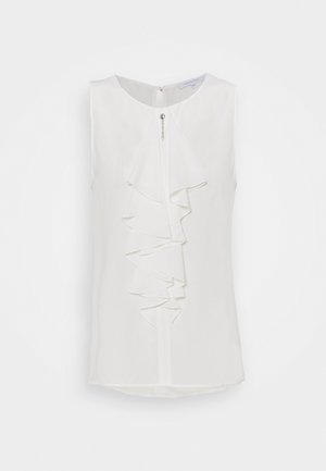 Blusa blanca sin mangas con volante delantero en capas y pequeño botón decorativo en el escote, mostrada sobre un fondo liso.