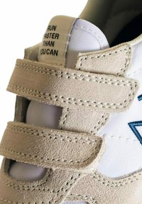 Sneaker di suede beige con parte superiore in mesh bianco, dotata di cinturini in Velcro e accenti ricamati blu. L'etichetta interna recita: "CORRI PIÙ VELOCE DI QUANTO PUOI."