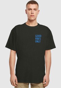 Zwarte katoenen t-shirt met korte mouwen, ronde hals en blauwe tekst met "GOEDE VIBRATIES ENKEL" aan de linkerkant.