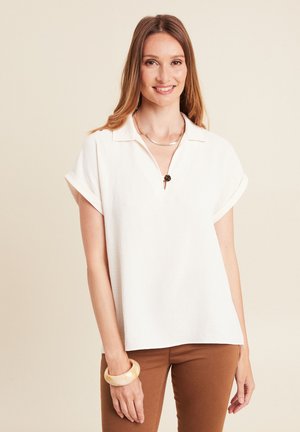 Vrouw met lang bruin haar, gekleed in een witte blouse met korte mouwen, een bruine broek, een gouden ketting en een beige armband, glimlachend tegen een effen achtergrond.